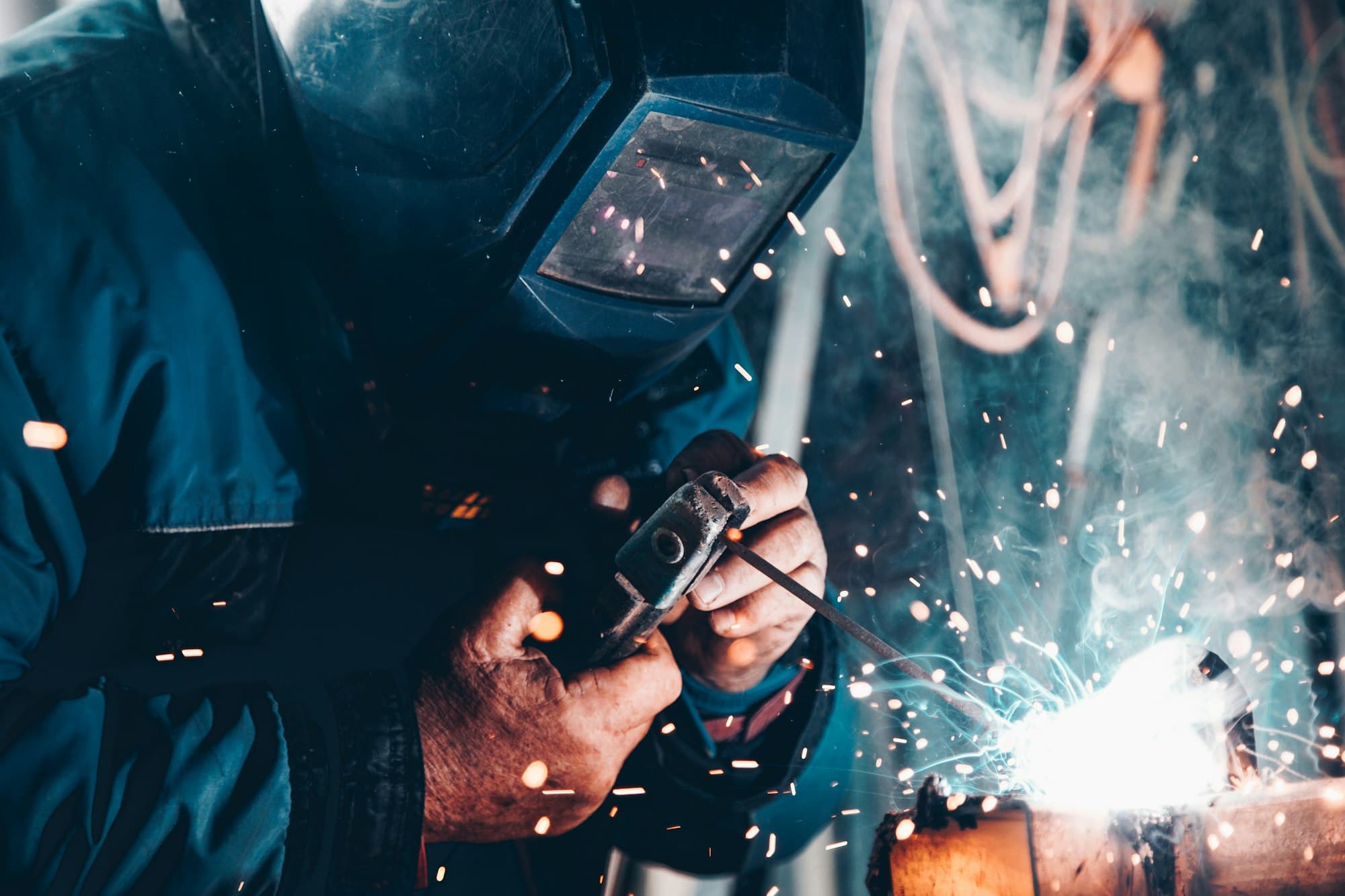 A welder in protective equipment joining a steel workpiece, with sparks visible from the arc.