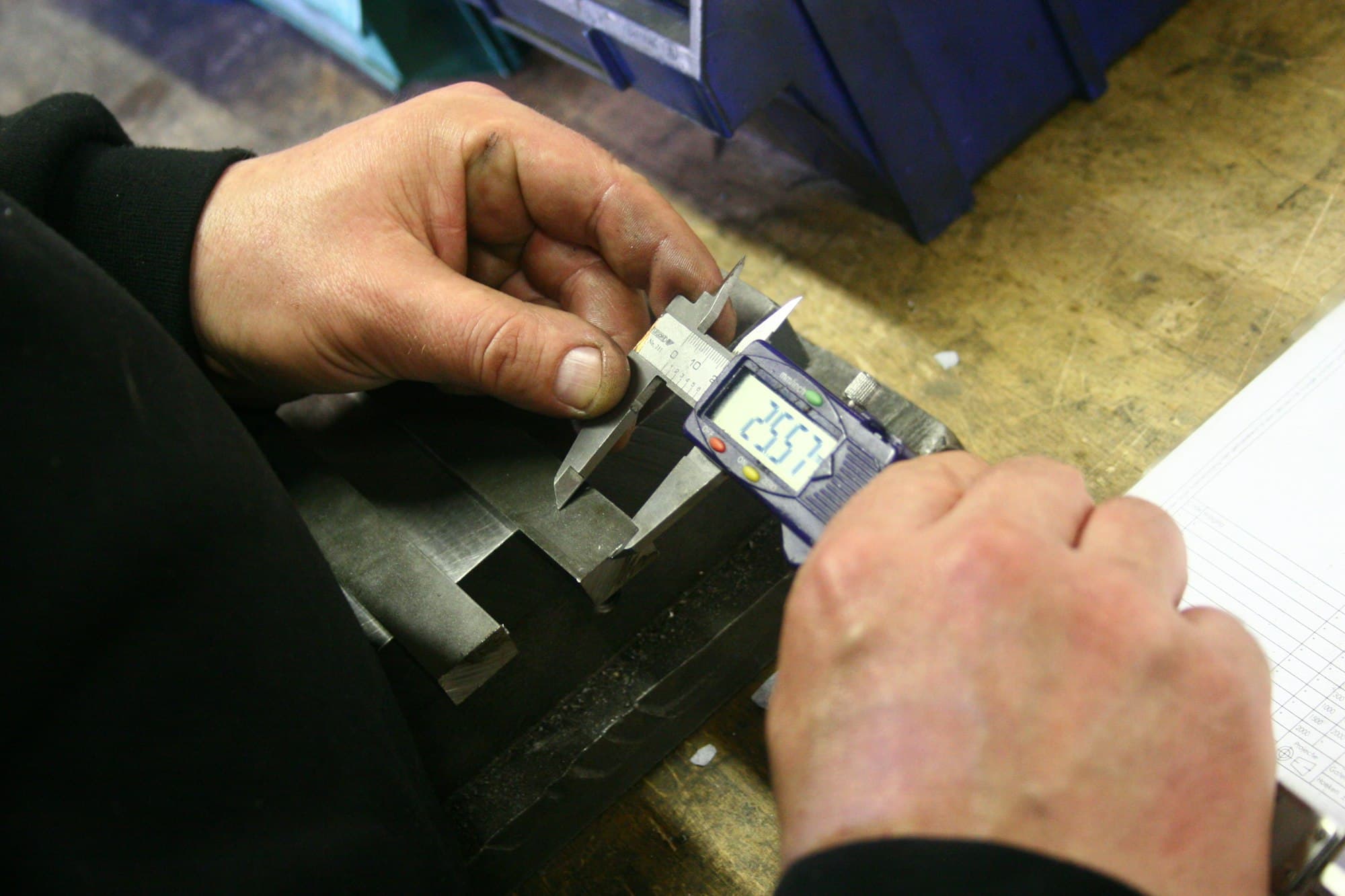 A technician using a digital caliper to check the dimensions of a machined metal part against a reference document.